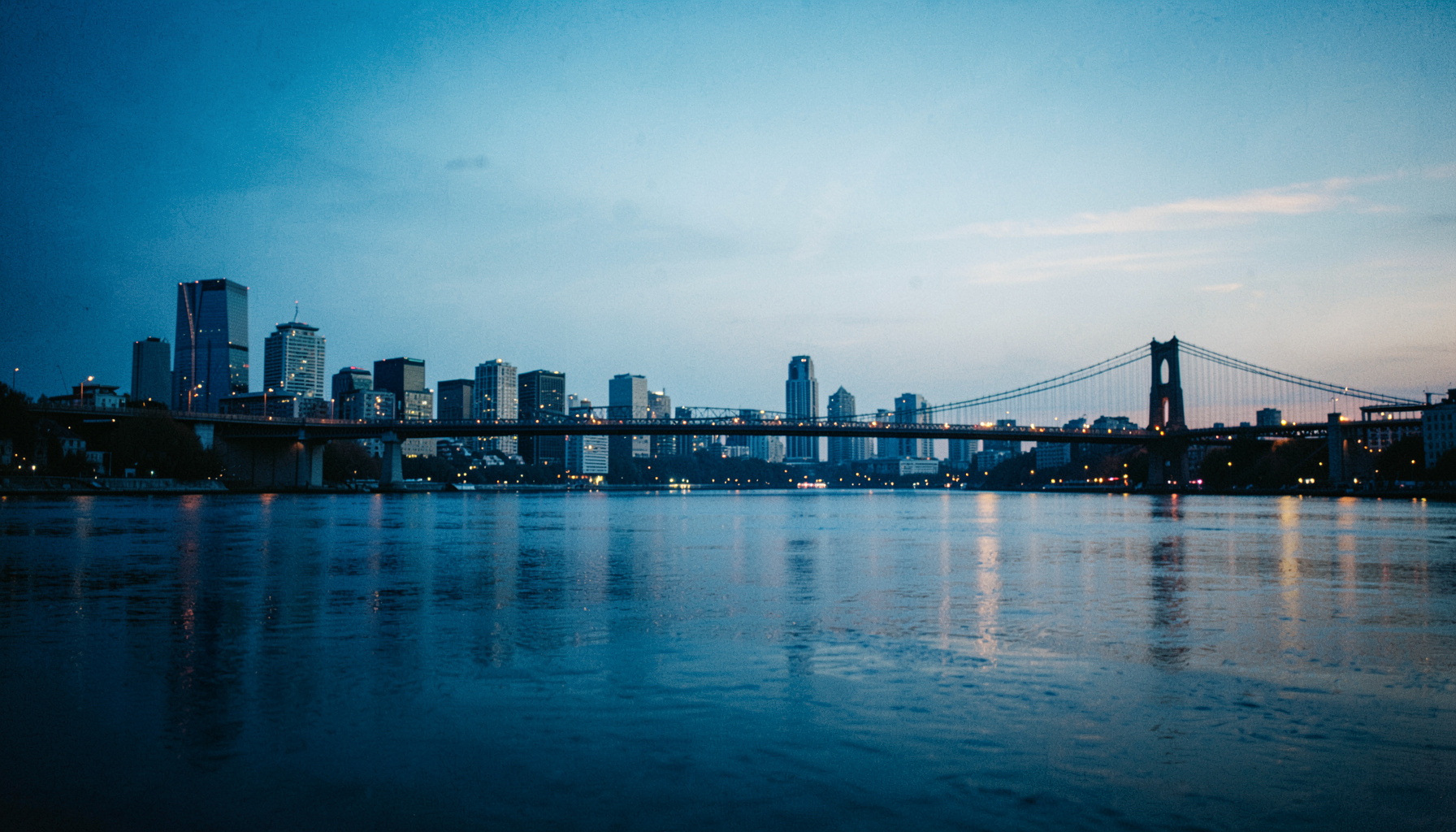 Wide cinematic view of a riverfront skyline at dusk with calm water in the foreground and bridges stretching across in soft blue and amber tones