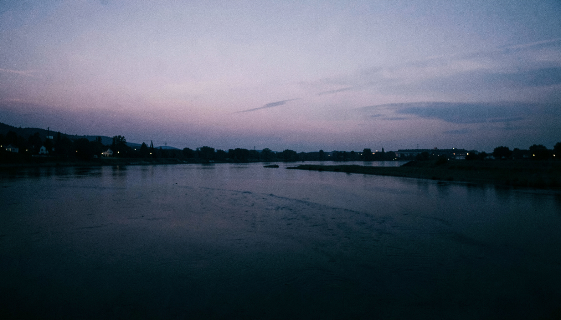 Panoramic riverfront view at dusk, soft purple and slate skyline fading into a calm dark river, atmospheric and empty.