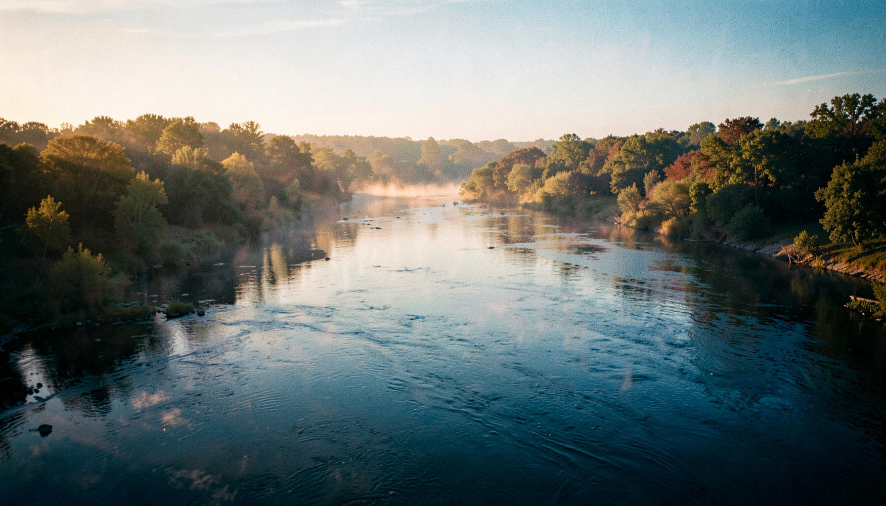Wide cinematic view of three rivers meeting at golden hour, soft haze over the water and tree-lined banks in muted golds, blues, and greens
