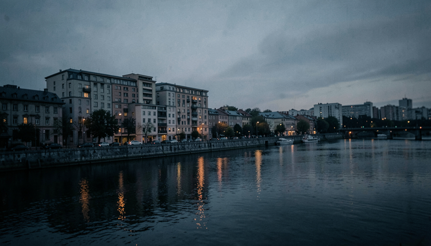 Wide cinematic view of a riverside city building along a calm river at dusk, muted charcoal gray and deep blue tones, soft warm light along the water's edge, shallow depth of field giving a quiet civic mood