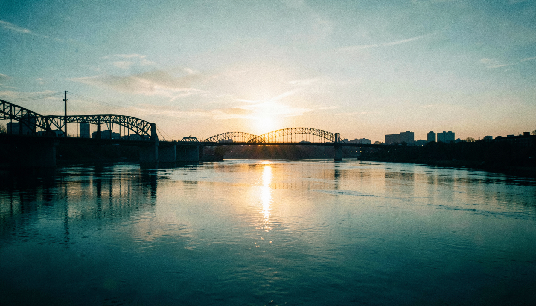 Wide cinematic riverscape of Pittsburgh with the sun rising behind silhouetted bridges, soft golden light reflecting on calm river surface, muted teal and amber tones.