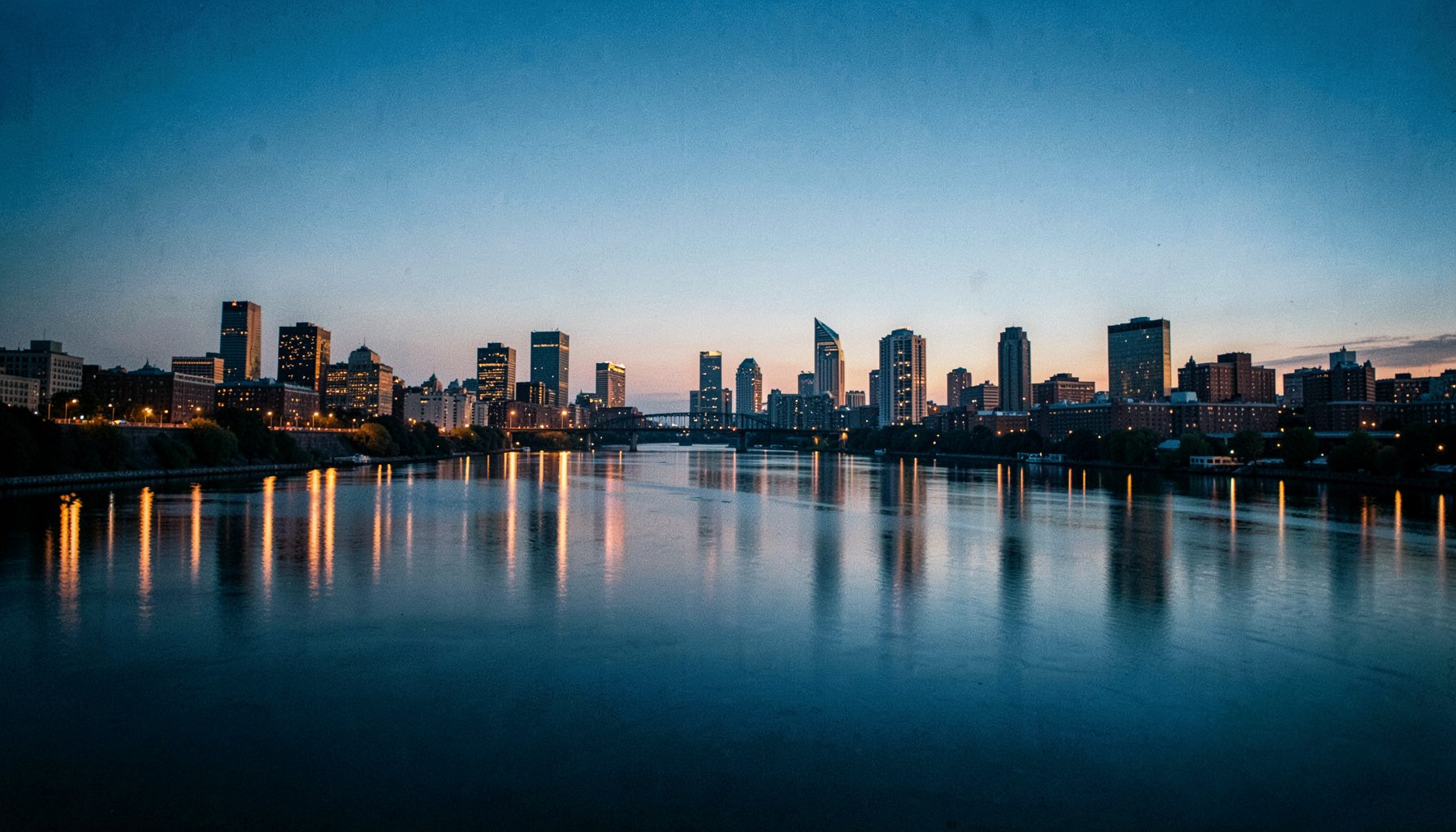 Wide cinematic view of Pittsburgh's three rivers at dusk with downtown skyline reflected in calm water, warm amber and cool steel blue tones